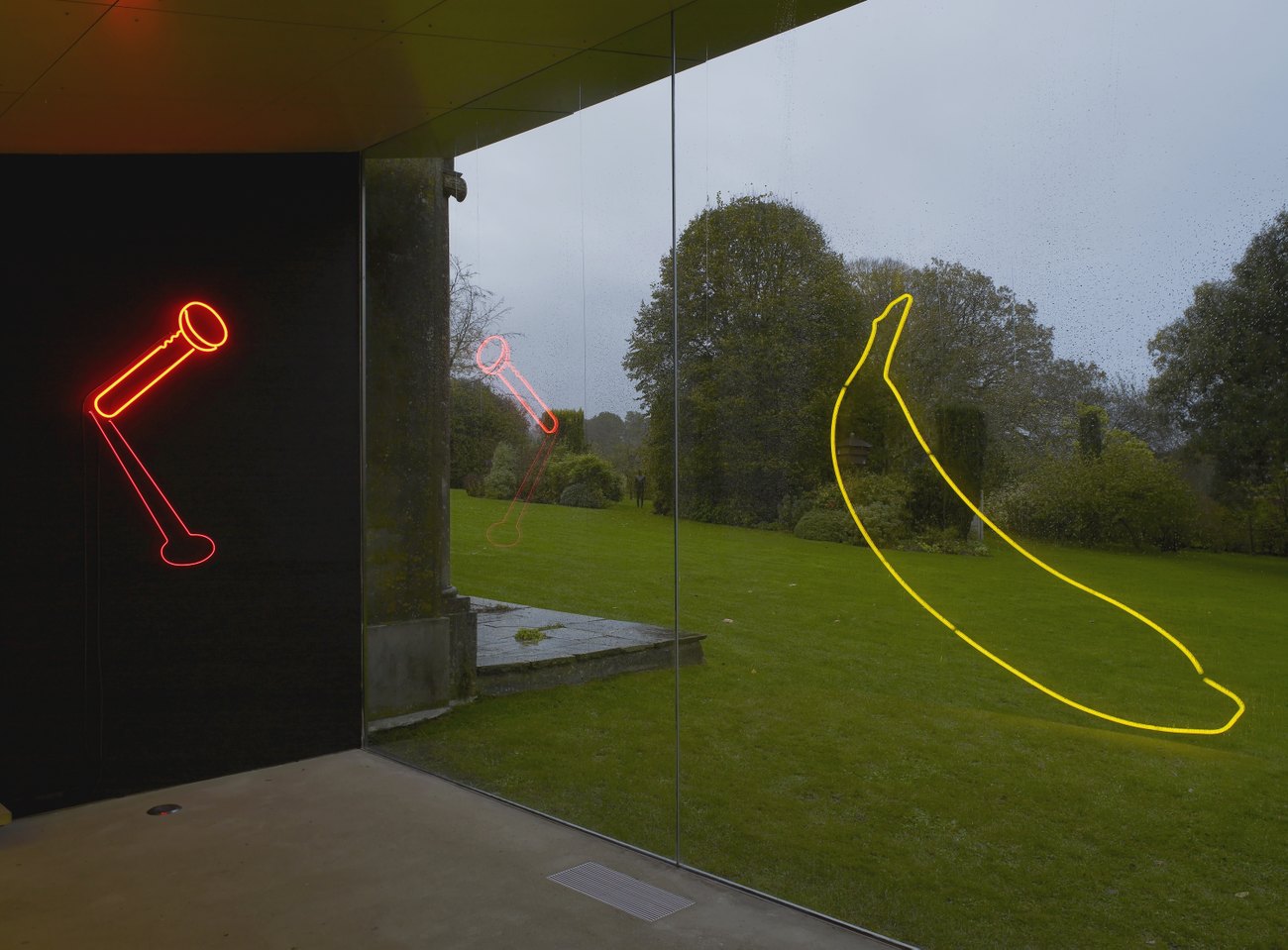 A red neon nail in the wall on a black backdrop, the reflection of a yellow neon banana reflected in a glass window overlooking a grassy garden
