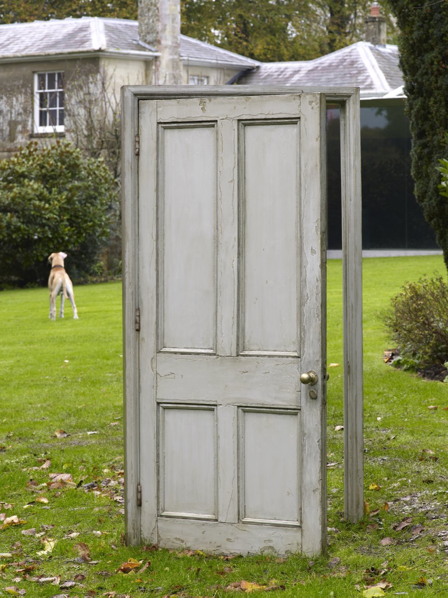 A sculpture of a freestanding doorway in a grassy garden with a dog and a house in the background