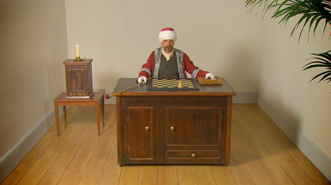 A mechanised figure sits at a wooden desk with a chess board before it, the figure is holding a long pipe. There is a small table to the left of the desk with a cabinet and a candle on top