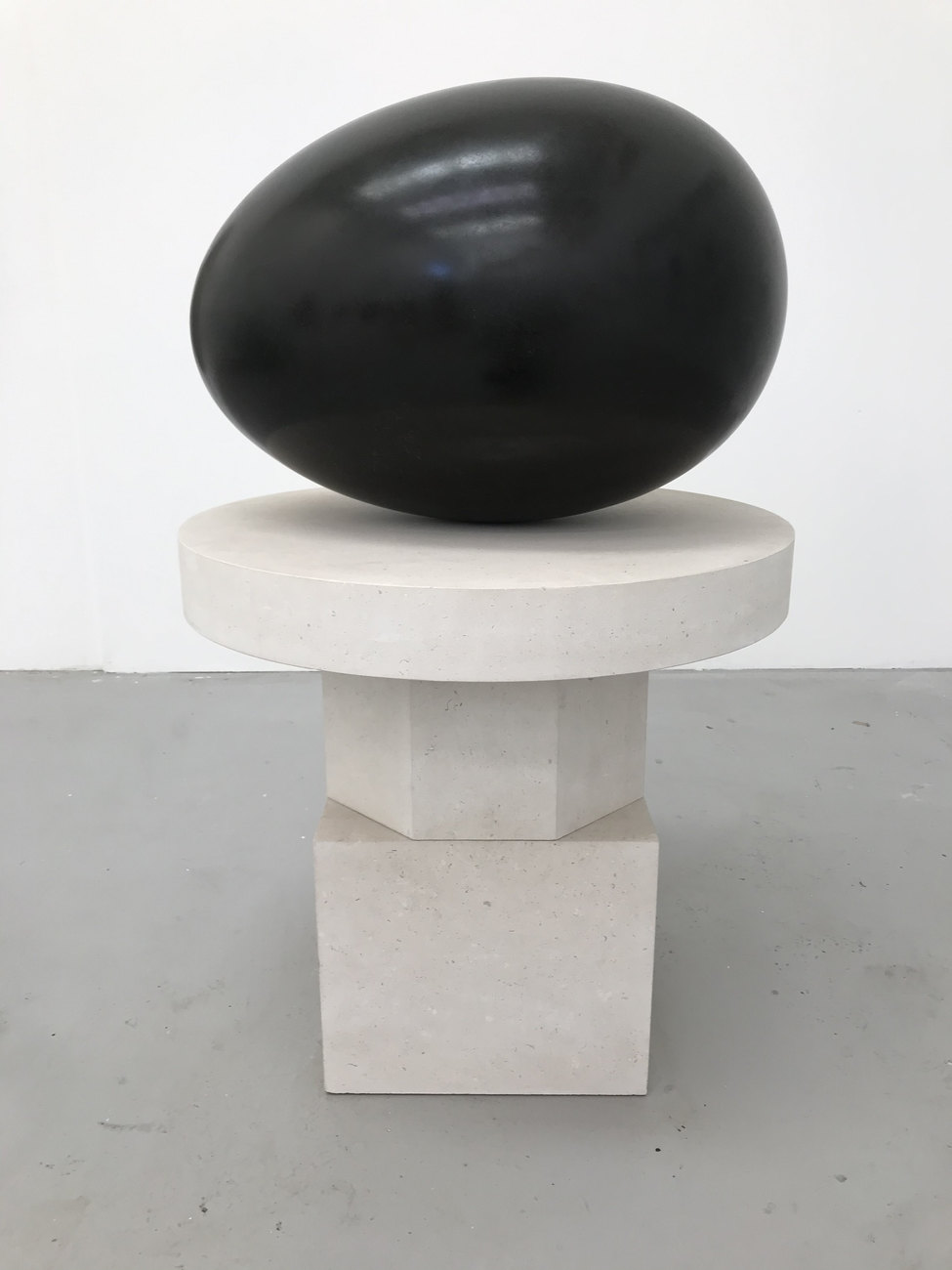 A large black egg sculpture on a stone plinth against a white walled background