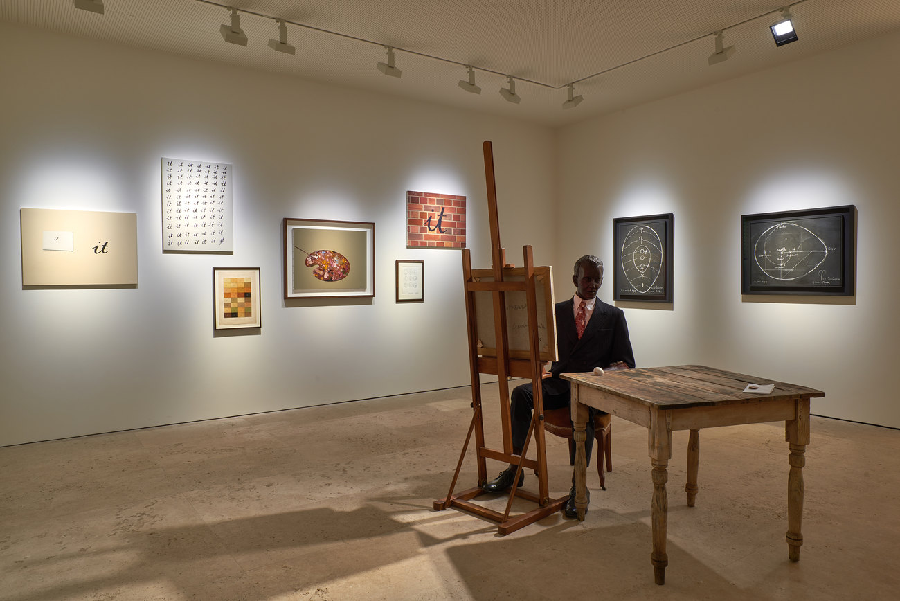 A sculpture of a man sits in the centre of a room with an easel in front of him and a table beside him with an egg on it. On the walls behind him are a series of paintings and images