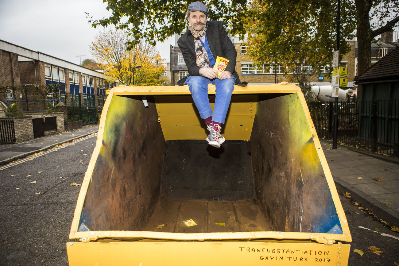 A man (Gavin Turk) sitting on a yellow skip, in a street, holding a bag of skips crisps