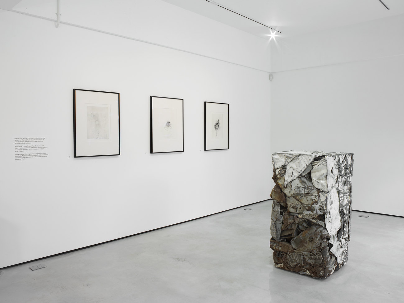 A white room with three framed etchings on the wall and a white van crushed into a cuboid shape in the middle of the room