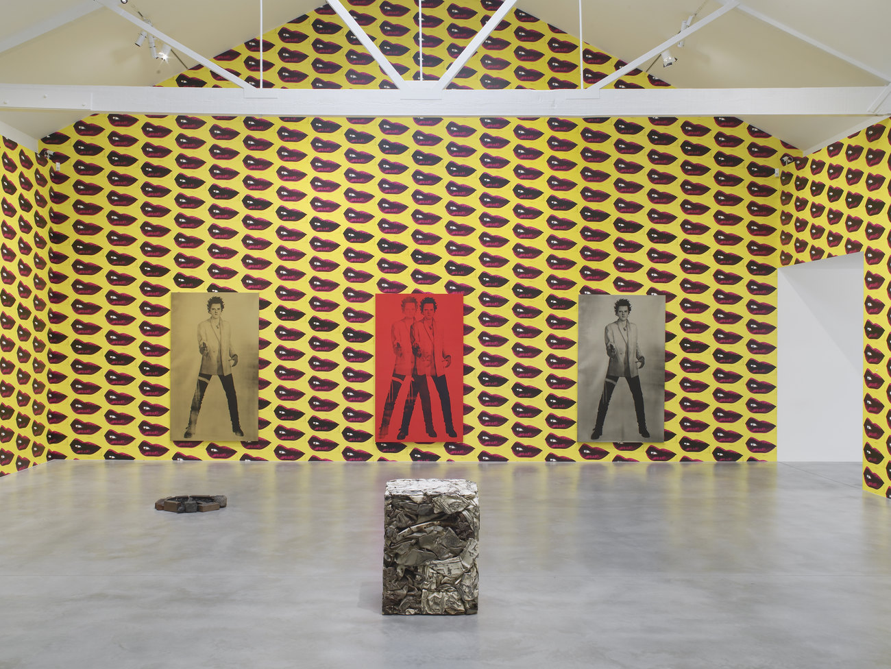 A compacted metal cuboid sits in the centre of a room, behind it hang three prints of a man (Gavin Turk) as Sid Vicious, adopting an iconic Elvis pose. The walls are yellow with red pattern