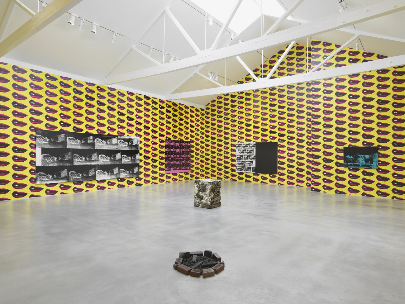 A compacted metal cuboid and a small brick firepit sit in the centre of a room, on the wall are four prints of different colours showing a white van crashing