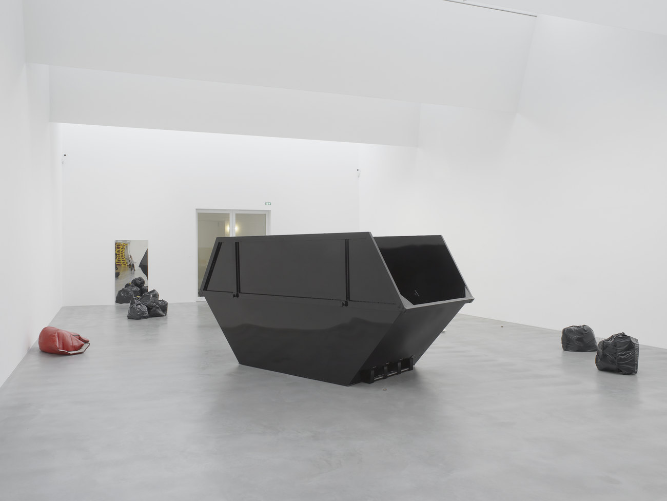 A large black skip site in the centre of a white room, around it are a few rubbish bags