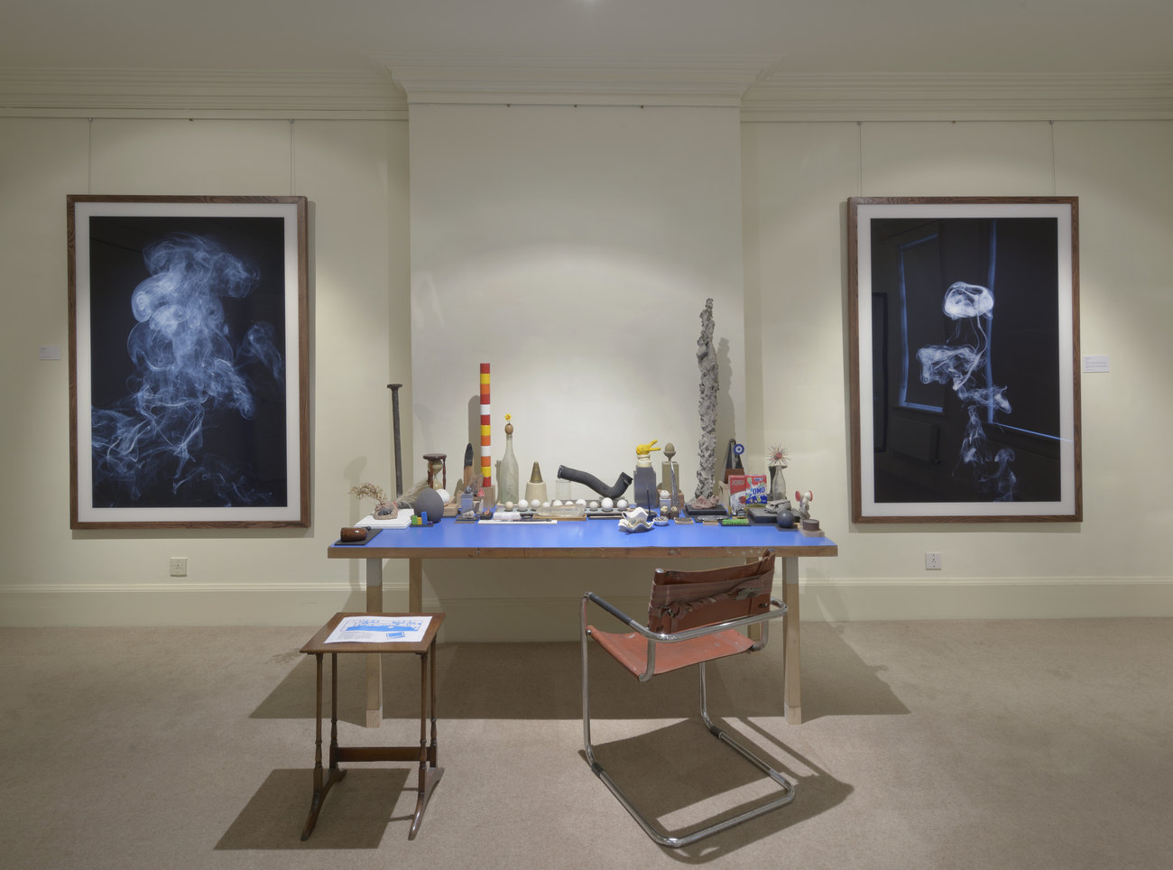 A blue desk covered with objects, many of them tall and long, with a red desk chair and a small table in front. On the white wall behind the desk are 2 large, framed photographs of billowing smoke against black backgrounds