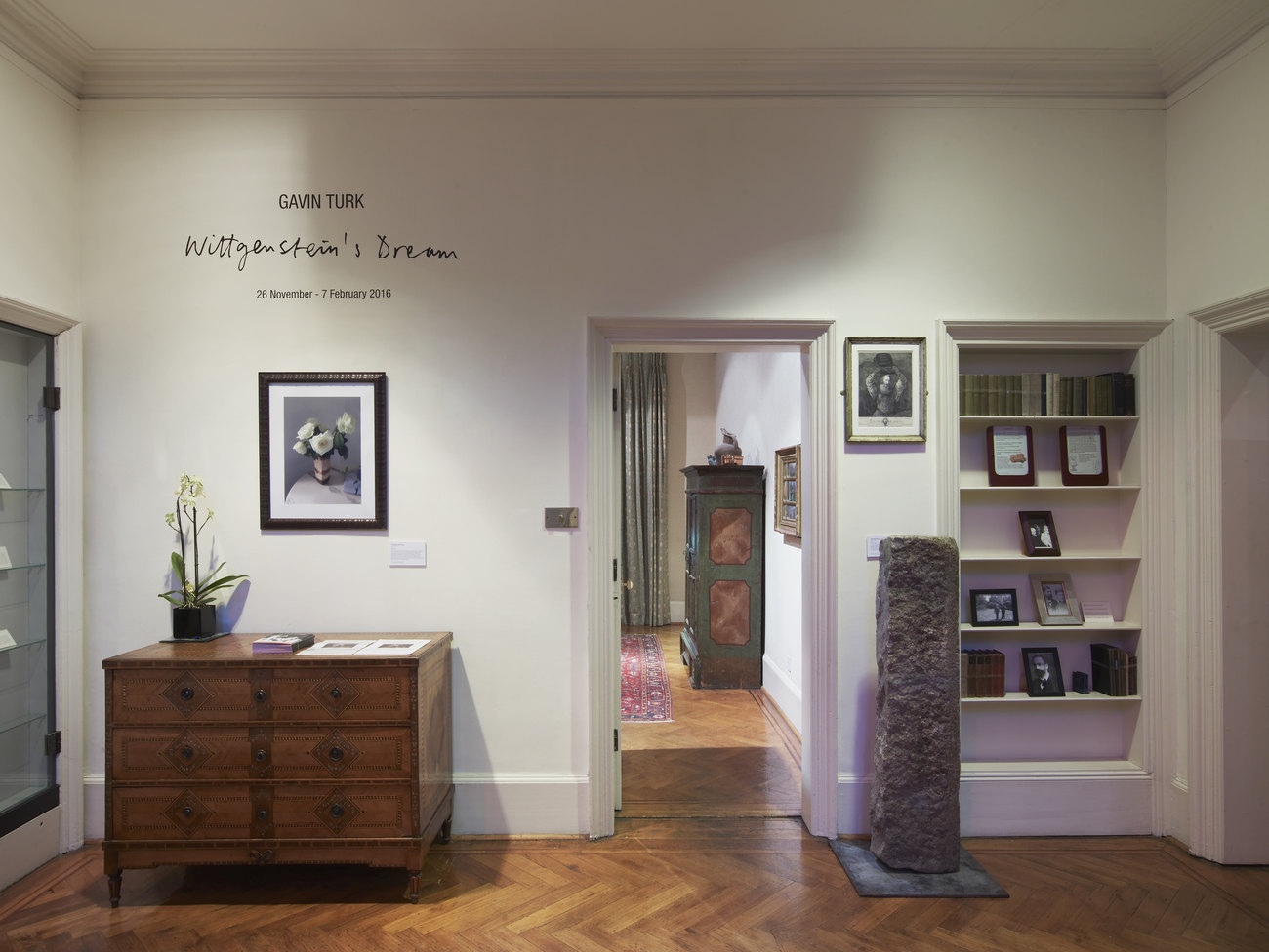 A room in The Freud Museum. There is a wooden set of drawers with a photograph of white flowers hung above to the left of a doorway. On the right of the doorway is a long stone column, roughly half the height of the doorway  