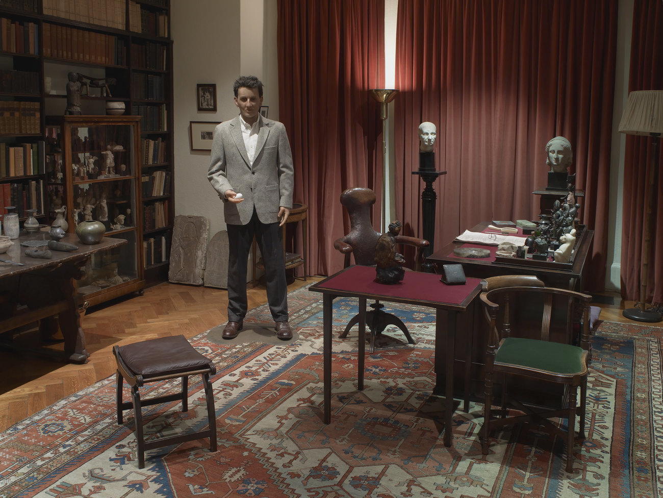 A waxwork of Wittgenstein holding an egg in The Freud Museum. It is stood in an office with a red rug, red curtains, a desk, some chairs and a book case against one wall