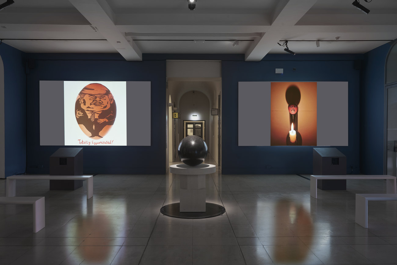 Installation photo of an exhibition incorporating projections of photos of artist's portraits of an egg, and Gavin Turk's sculpture Oeuvre (Black Umber) 