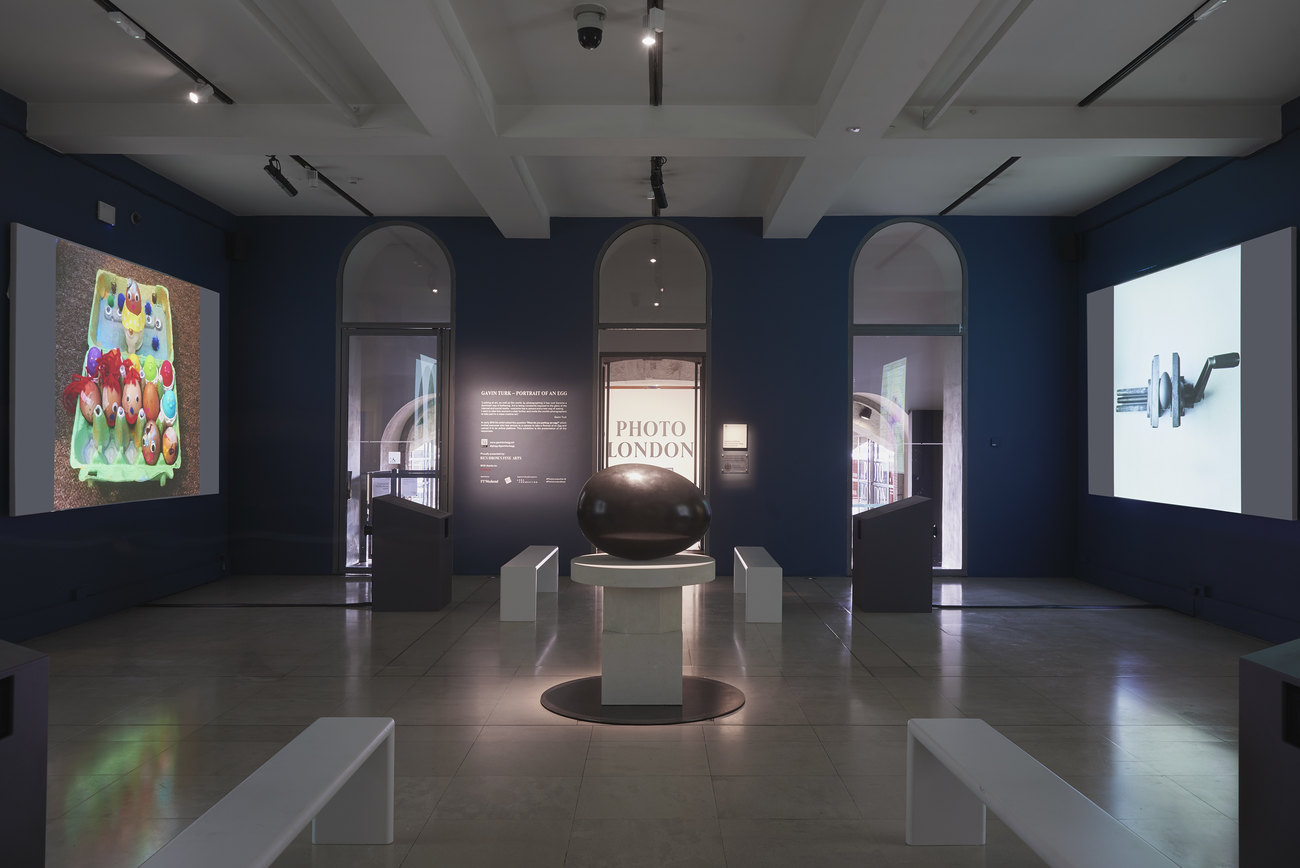 Installation photo of an exhibition incorporating projections of photos of artist's portraits of an egg, and Gavin Turk's sculpture Oeuvre (Black Umber) 