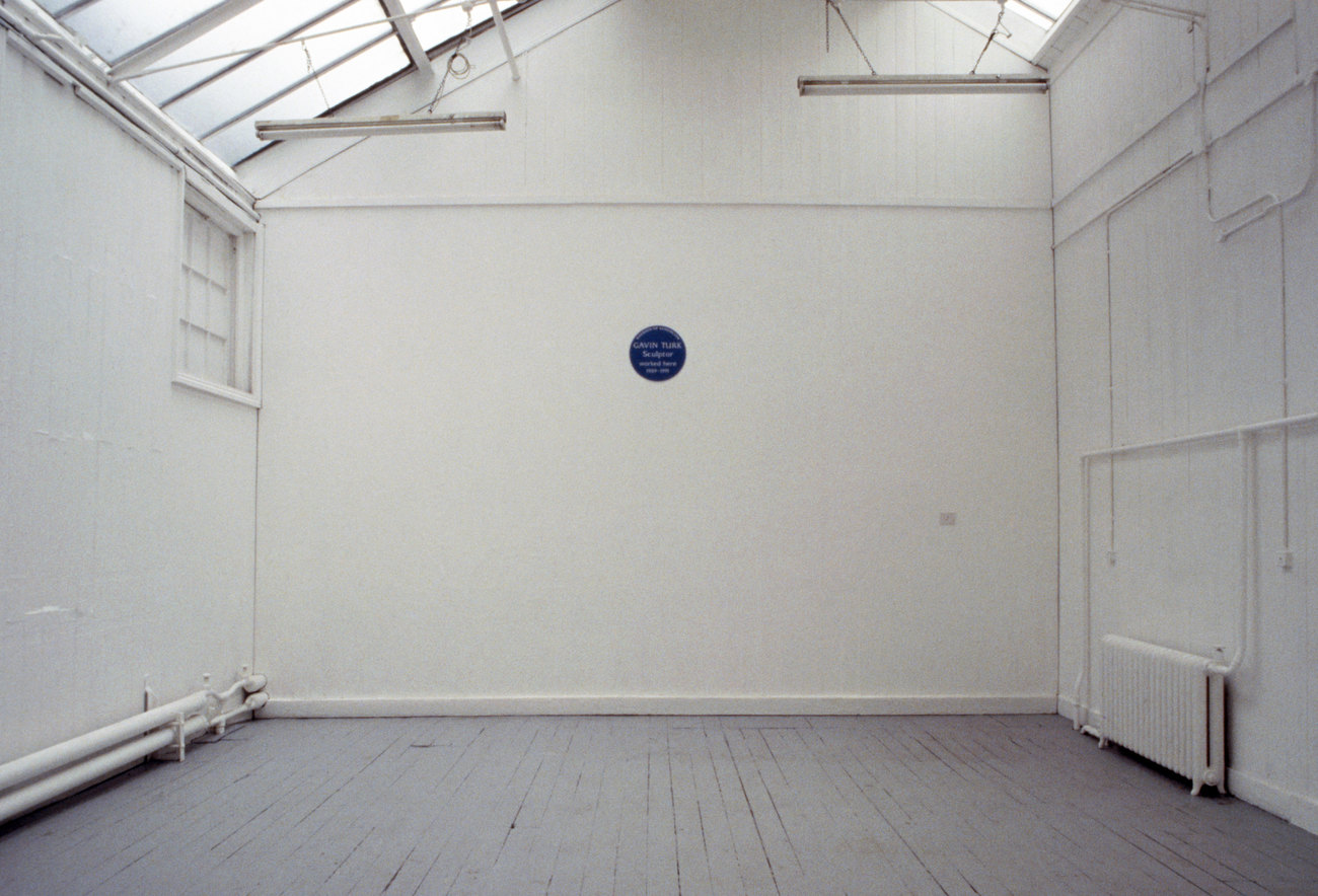 A white walled space is empty, expect a blue english heritage plaque showing the words Gavin Turk Sculptured Here 
