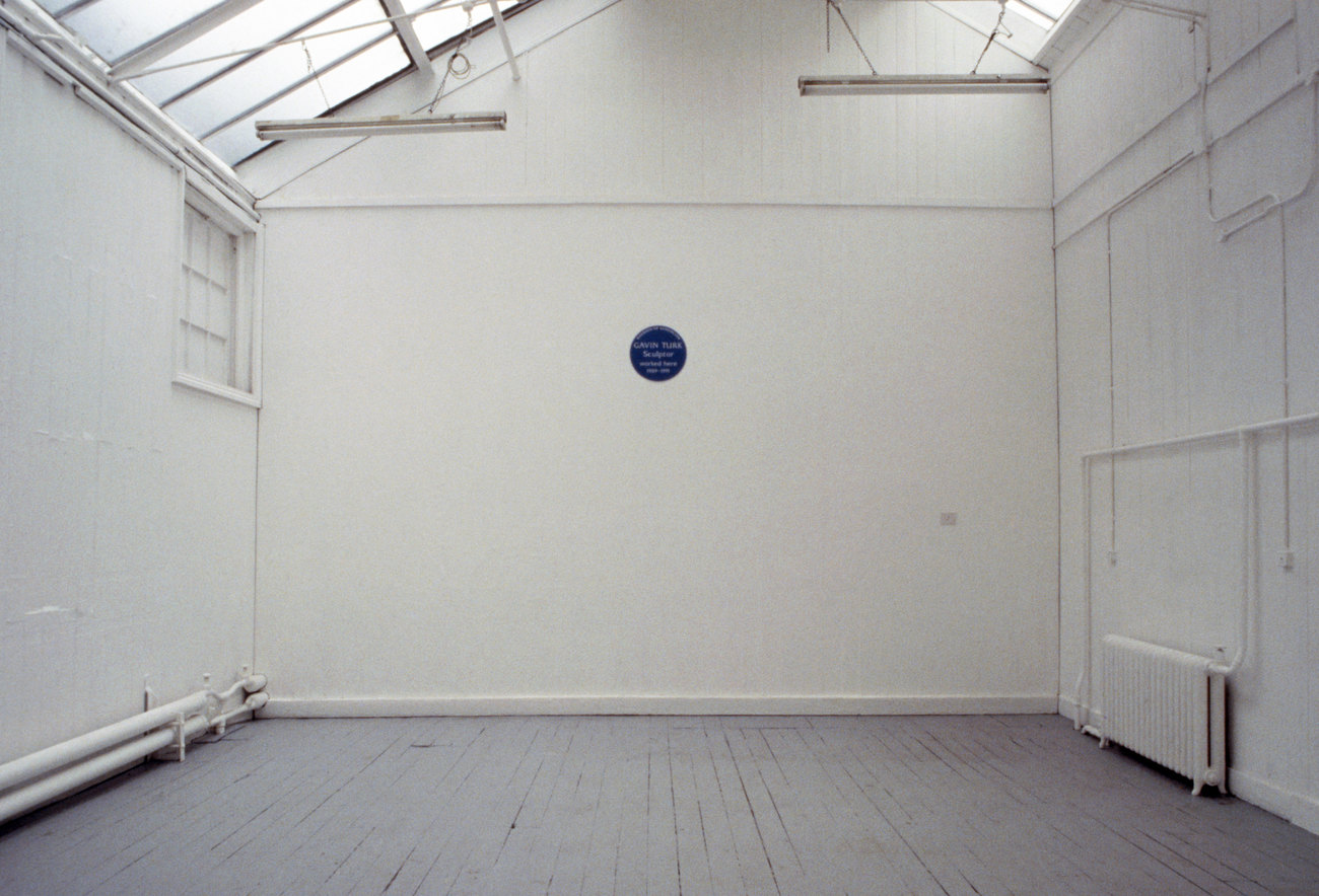 A white walled space is empty, expect a blue english heritage plaque showing the words Gavin Turk Sculptured Here