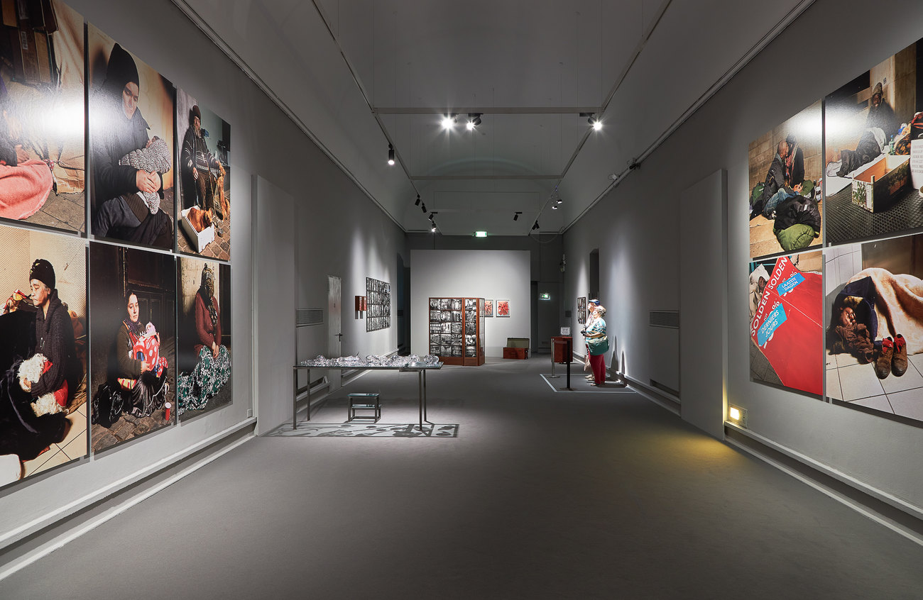 Installation photo of a group exhibition, on the walls are photos of homeless people, within two figures appearing as American tourists look at imagery, a table is filled with glass ware 