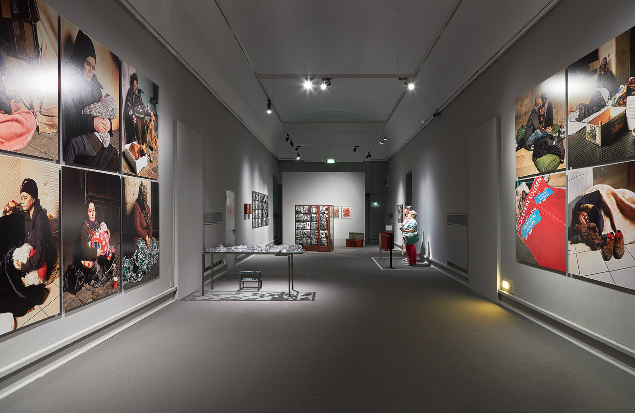 Installation photo of a group exhibition, on the walls are photos of homeless people, within two figures appearing as American tourists look at imagery, a table is filled with glass ware