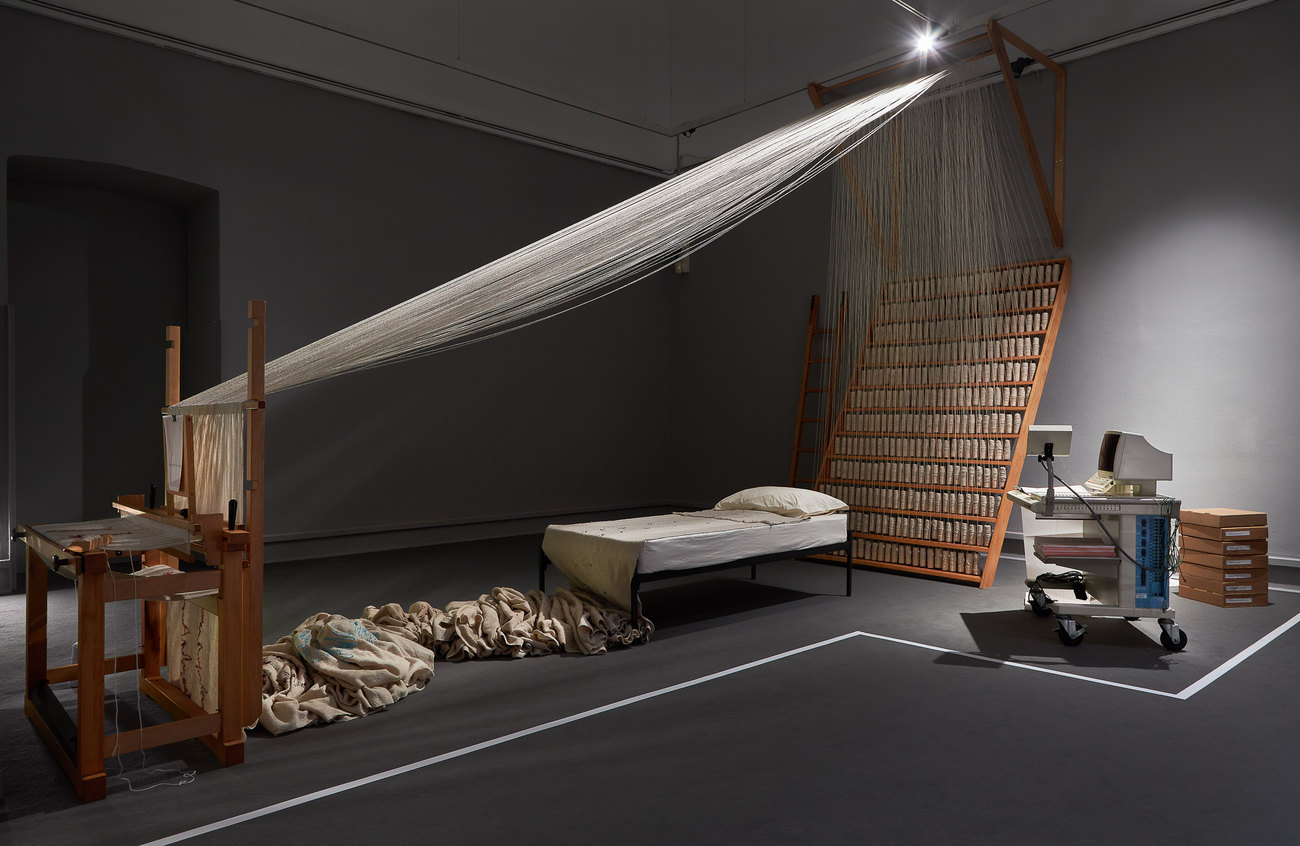 an installation photo of an exhibition shows a weave machine, the weaved cloth is then turned into a bed spread, a bed with what appears as hospital machinery sits within