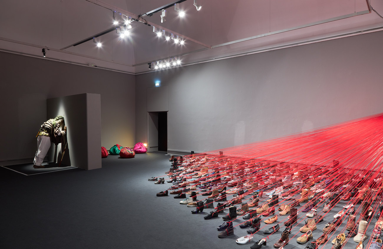 installation photo of an exhibition shows, red thread attached to approx 100 shoes pointing away from the direction of the wall. And a fugure with his head on the wall, lit from above