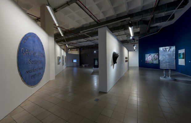 A picture shows an installation photo on the wall is a rug, the rug resembles a large Blue Plaque with the words Gavin Turk sculptured here.