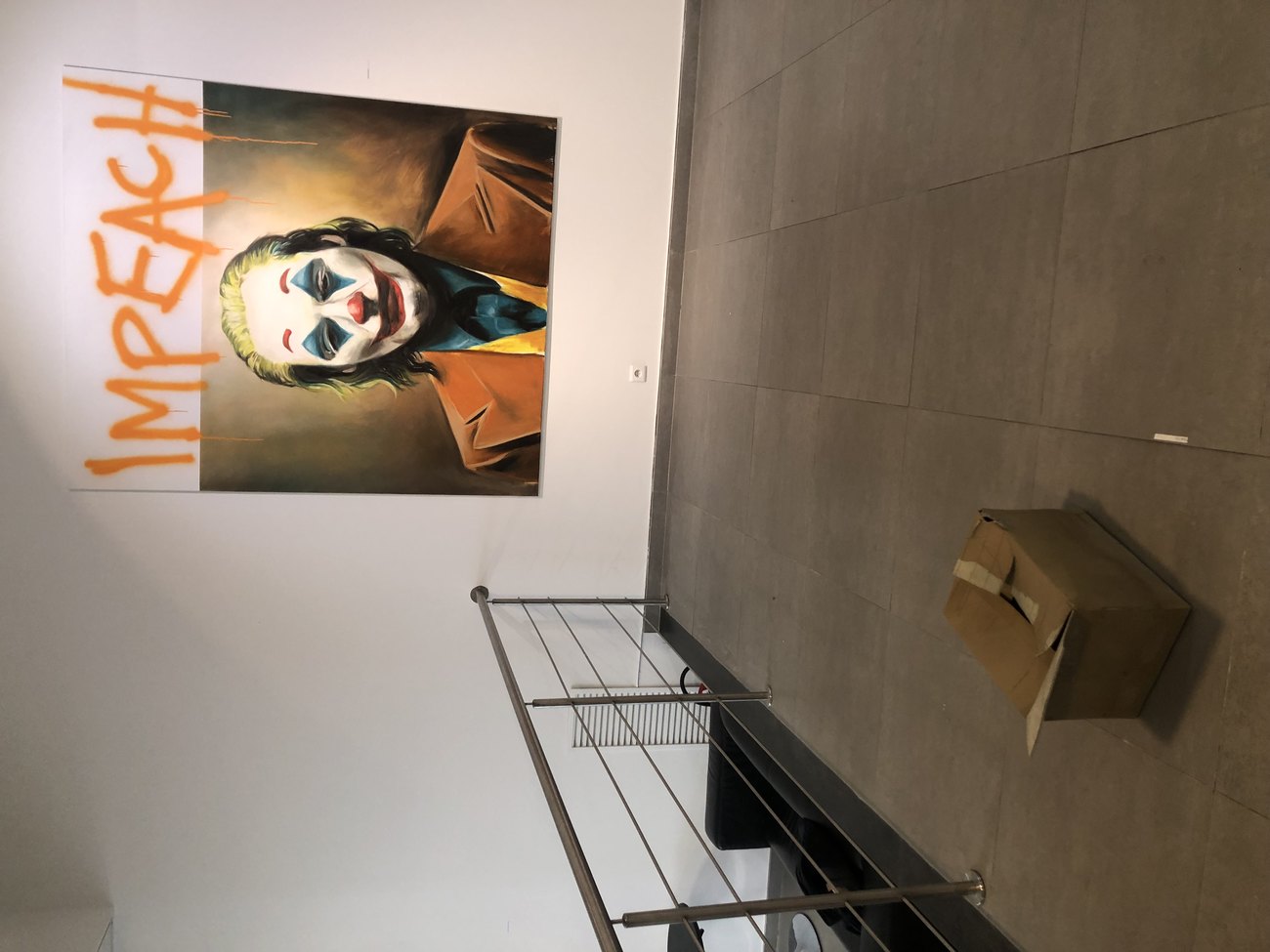 An image shows a painting of the joker with IMPEACH spraypainted on top, and a cardboard box made of bronze on the floor