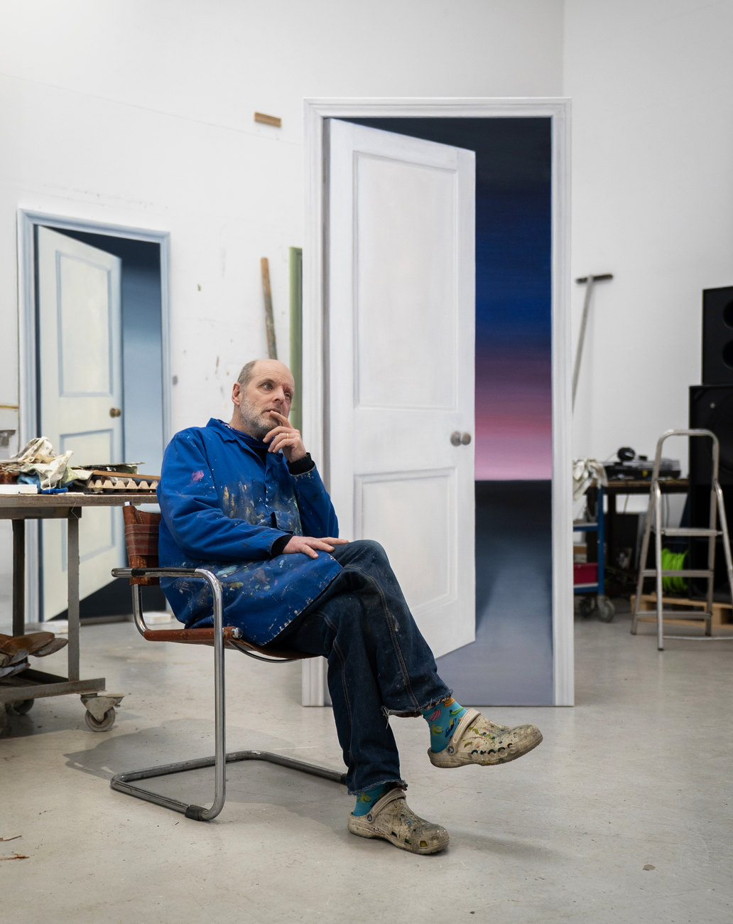Image shows the Artist Gavin Turk sat on a chair in front of painting of a door set ajar.